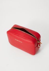 Raudona odinė tualetinių reikmenų dėžutė su tekstūruota paviršiumi, auksinėmis užtrauktukų detalėmis ir furnitūra. Priekyje yra "VALENTINO" logotipas. Stačiakampio formos.