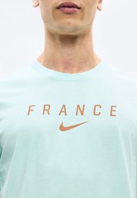 Άνδρας που φοράει ανοιχτό μπλε πουκάμισο με το λόγκο "FRANCE" και το πορτοκαλί λογότυπο Nike στο στήθος, με φόντο απλό λευκό.