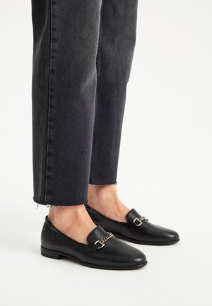 Czarne skórzane mokasyny z dodatkiem złotego łańcuszka, w połączeniu z przyciętymi, ciemnoszarymi dżinsami z postrzepionymi końcami.