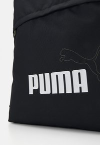 Svart stoffryggsekk med et stort hvitt "PUMA"-logo og en gylden silhuett av en puma. Glidelåsdetaill ved toppen.
