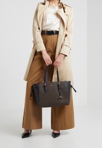 Zwarte en bruine tote-handtas met een gestructureerd oppervlak, gouden hardware en een logo charme. De tas heeft dubbele handvatten en een hoekige vorm.