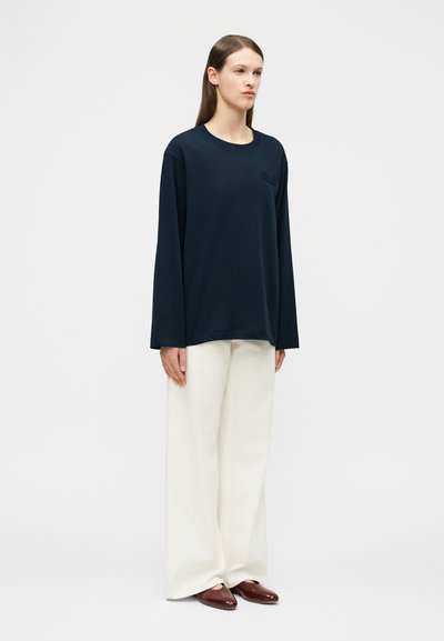 Γυναίκα που στέκεται, φοράει μπλε σκούρο μακρυμάνικο πουκάμισο, λευκό παντελόνι με φαρδιά πόδια και καφέ επίπεδα παπούτσια, μπροστά σε απλό φωτεινό φόντο.