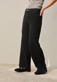 Sorte viddejeans lavet af denim med høj talje, forlommer og diskrete syninger. Kombineret med sorte slip-on sko.