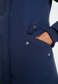 Navy blue jacket featuring a smooth, textured fabric, side pockets with snap closure, and ribbed cuffs.