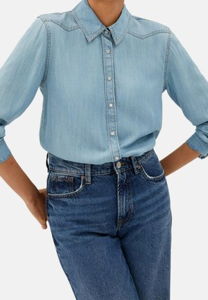 Personne portant une chemise en jean bleu clair à boutons avec col, rentrée dans un jean taille haute bleu foncé, les bras pliés sur les côtés.