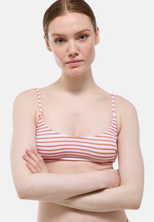 Jeune femme à la peau claire et aux cheveux roux portant un haut de bikini rayé orange et blanc, bras croisés, sur un fond blanc uni.