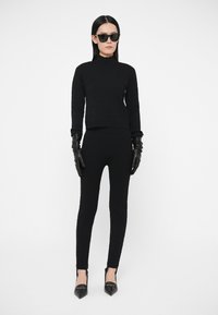 Pulover negru tricotat cu textură striată, asortat cu pantaloni negri mulați. Modelul poartă mănuși negre din piele și ochelari de soare negri, cu unghiuri ascuțite.