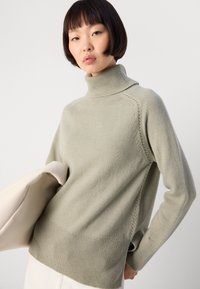 Young woman with short dark hair wearing a light gray turtleneck sweater and holding a beige bag against a plain background.