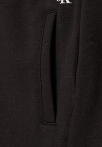 Czarne dresy bawełniane z boczną kieszenią i widocznymi szwami. Faktura wygląda na gładką i miękką. Biały nadruk z logo na przodzie.