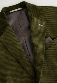 Olivazöld corduroy blazer texturált anyaggal, bemetszett gallérral és sötét mintás bélésnel. Mellzsebet és diszkrét fém részleteket tartalmaz.