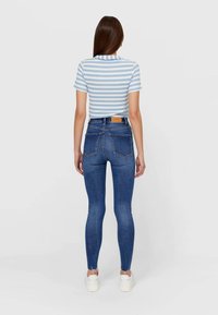 Mujer de pie con la espalda hacia la cámara, vistiendo una camisa de rayas azul claro y blanca de manga corta, jeans ajustados azul de cintura alta y zapatillas blancas.