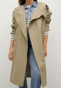 Trench-coat beige avec grands revers, manches longues, et fermetures à pression. Porté sur une veste en jean et un haut rayé, associé à un jean bleu.