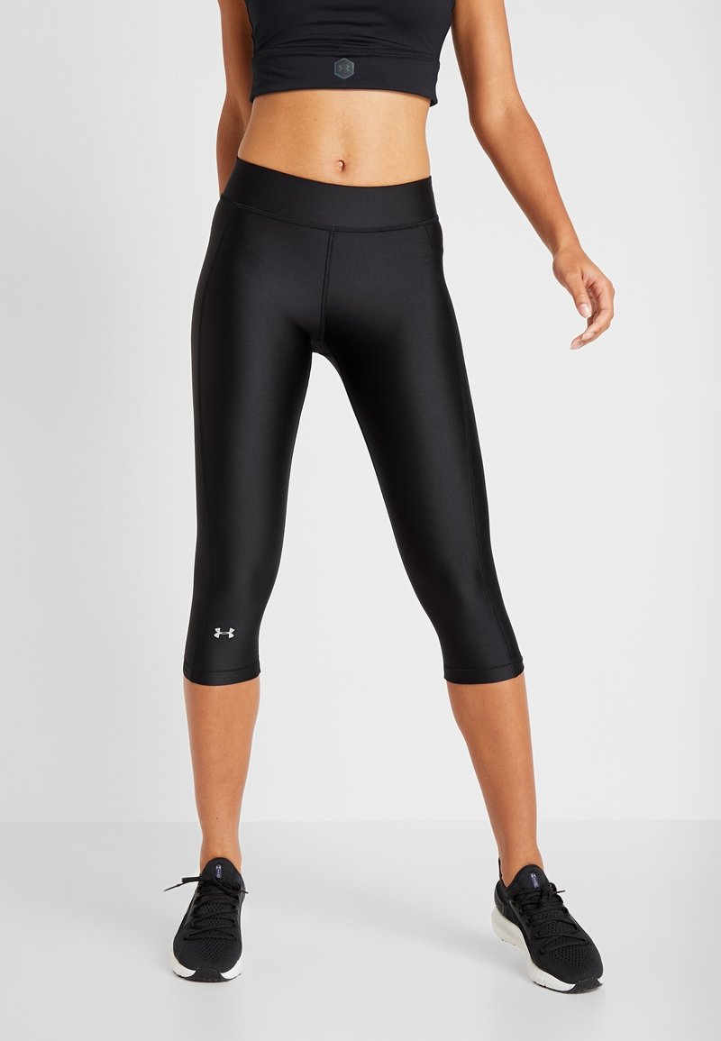 Femme portant des leggings noirs courts, un soutien-gorge de sport noir et des chaussures de sport noires sur un fond uni.