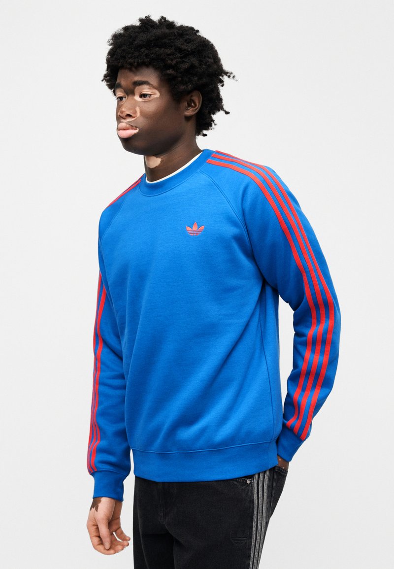 Sweat-shirt bleu en coton, avec des détails à trois bandes rouges sur les manches et un petit logo rouge sur la poitrine. Col rond.