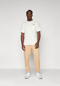 Ανοιχτό γκρι βαμβακερό μπλουζάκι με τσέπη στο στήθος και μαύρη εκτύπωση, συνδυασμένο με μπεζ στενά φαρδιά φόρμες και λευκά αθλητικά παπούτσια.