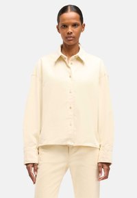 Camicia oversize color crema con chiusura a bottoni, in morbido cotone, colletto a punta e maniche lunghe, abbinata a pantaloni coordinati.