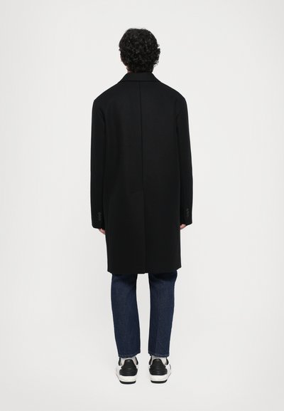 Long black coat with a straight silhouette and notched collar, paired with blue jeans and black sneakers. Features buttoned sleeves.
