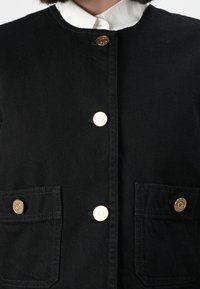 Veste noire en toile avec un col rond, des boutons dorés et deux poches avant.