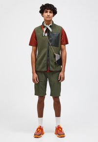 Green fleece vest with black pocket, red t-shirt, olive shorts, white socks, and orange sneakers. Accessories include a patterned scarf and small bag.