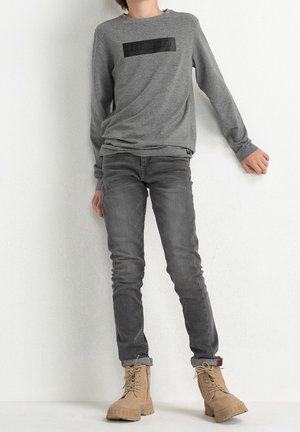 Person, der læner sig op ad en hvid væg iført en grå langærmet skjorte, mørkegrå jeans og beige snørestøvler.
