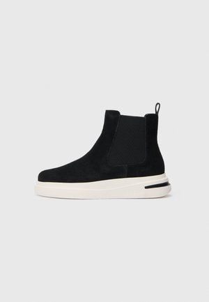 Stivale Chelsea in suede nero con pannelli elastici laterali e suola di gomma bianca con una striscia nera sul tallone.