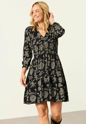 Blonde Frau lächelnd, trägt ein schwarzes Kleid mit beigen Blumenmustern, langen Ärmeln und enger Taille, posiert vor hellem Hintergrund.
