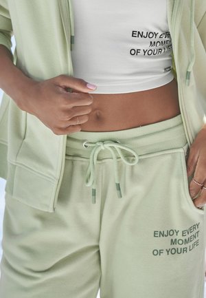 Hellgrünes sportliches Outfit, bestehend aus einem Reißverschluss-Hoodie und Jogginghosen. Das Oberteil hat ein weißes Crop-Design, während die Hose einen aufgedruckten Spruch zeigt.