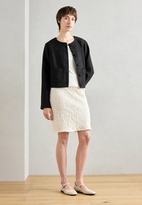 Svart teksturert kort jakke med knappelukking og to frontlommer, kombinert med et hvitt krøllete skjørt og lysebeige slingback-sko.