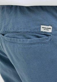 Jasnoniebieskie materiałowe spodnie z tylną kieszenią i małą białą metką z napisem "JACK & JONES EST.1990". Gładka tekstura.