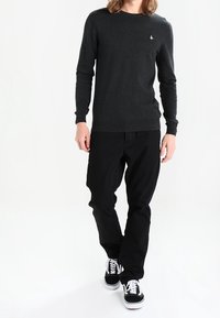 Grafitowy sweter z okrągłym dekoltem w zestawieniu z czarnymi spodniami i czarnymi sneakersami. Sweter ma małe białe logo na piersi.