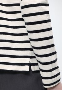 Λευκή και μαύρη μακρυμάνικη μπλούζα με οριζόντιες ρίγες και μικρό πλαϊνό σκίσιμο στο τελείωμα, φορεμένη πάνω από σκούρο παντελόνι.