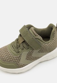 Zapatilla deportiva de malla verde con cordones, una correa de velcro, cuello acolchado y suela blanca etiquetada "AEROTECH" sobre un fondo liso.