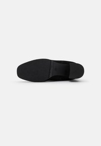Semelle de chaussure en daim noir avec une surface texturée. Dispose d'un bout arrondi et d'un talon légèrement surélevé. Design minimaliste sans quincaillerie visible.