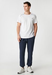 Biała koszulka sportowa z krótkim rękawem, z szarymi akcentami stębnowania, zestawiona z granatowymi spodniami dresowymi i białymi trampkami. Prosty, funkcjonalny design.