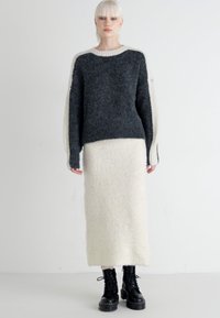 Maglione grigio e crema a maglia con maniche lunghe, abbinato a una gonna bianca, morbida, alla caviglia. Stivali neri con lacci completano l'outfit.