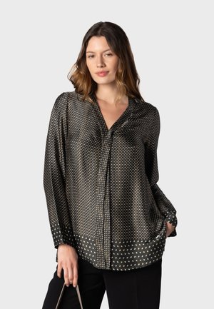 Femme portant un chemisier à manches longues avec motifs foncés et col, partiellement rentré dans un pantalon noir, tenant un sac dans une main.