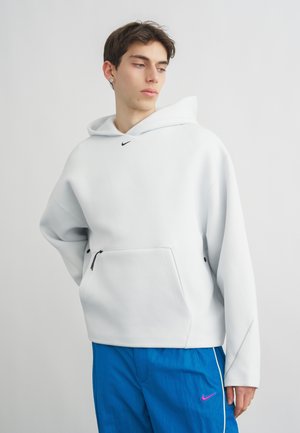 Jaunas vyras dėvi baltą Nike džemperį su priekine užtrauktuko kišene ir mėlynas Nike sportines kelnes, fone paprasta šviesi siena.