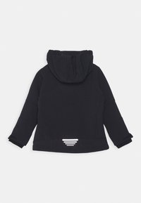 Veste à capuche noire avec des manches longues, arborant un accent réfléchissant et un tissu texturé. Le design est simple et ajusté, idéal pour superposer.