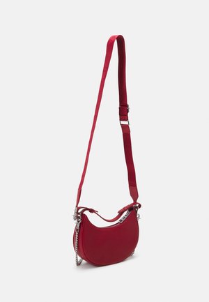Sac bandoulière rouge en forme de croissant avec sangle réglable et fermeture éclair argentée avec détail chaîne sur fond uni.
