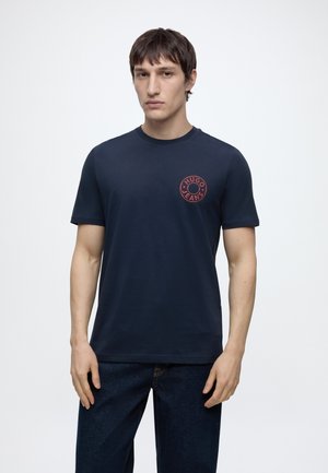 Άνδρας που φοράει σκούρο μπλε T-shirt με μικρό κόκκινο στρογγυλό λογότυπο "Hugo Jeans" στο στήθος, συνδυασμένο με σκουρομπλε τζιν.