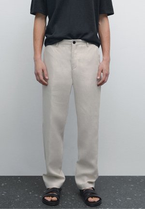 Homme portant un pantalon beige, un t-shirt noir et des sandales noires debout sur un sol sombre tacheté contre un mur uni gris clair.
