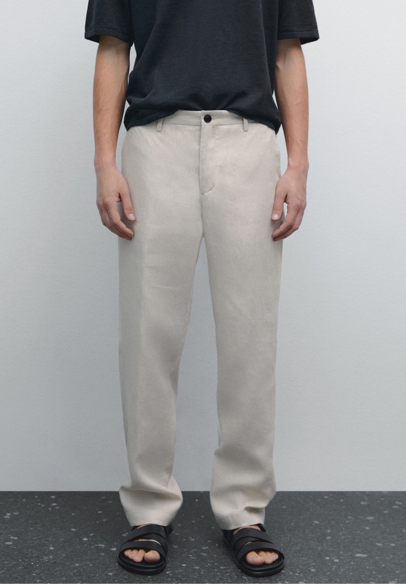 Homme portant un pantalon beige, un t-shirt noir et des sandales noires debout sur un sol sombre tacheté contre un mur uni gris clair.