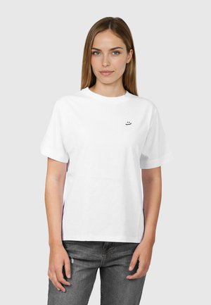 Junge Frau mit glattem hellbraunem Haar trägt ein weißes kurzärmeliges T-Shirt mit kleinem schwarzem Smiley-Logo auf der Brust, kombiniert mit grauen Jeans.