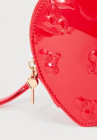 Κόκκινη τσάντα σε σχήμα καρδιάς από γυαλιστερό υλικό με ανάγλυφα σχέδια κερασιών. Περιλαμβάνει χρυσό κλειδί και φερμουάρ σε σχήμα καρδιάς.