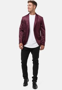 Blazer bordeaux in tessuto liscio, chiusura a un bottone, due tasche frontali, indossato sopra una camicia bianca, abbinato a jeans neri e scarpe.