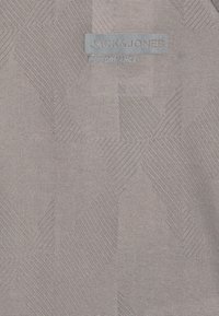 Grått tyg med ett strukturerat geometriskt mönster, prytt med "Jack & Jones Performance"-logotypen i en subtil blå etikett.