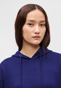 Námornícky modrá mikina s kapucňou s textúrovaným povrchom, ktorá má kovové očká a diskrétny logotyp značky na hrudi.