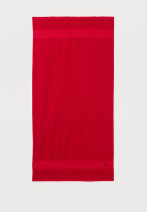 Serviette rectangulaire rouge vif avec des bandes bordures étroites en haut et en bas, petit logo brodé bleu foncé près du bord inférieur.