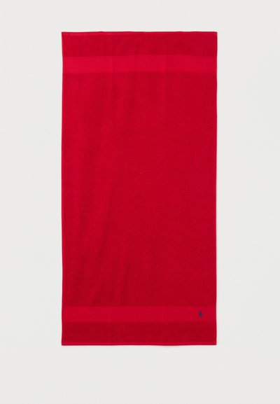 Lyse rød rektangulært håndklæde med smalle kantstriber øverst og nederst, lille mørkeblå broderet logo nær nederste kant.