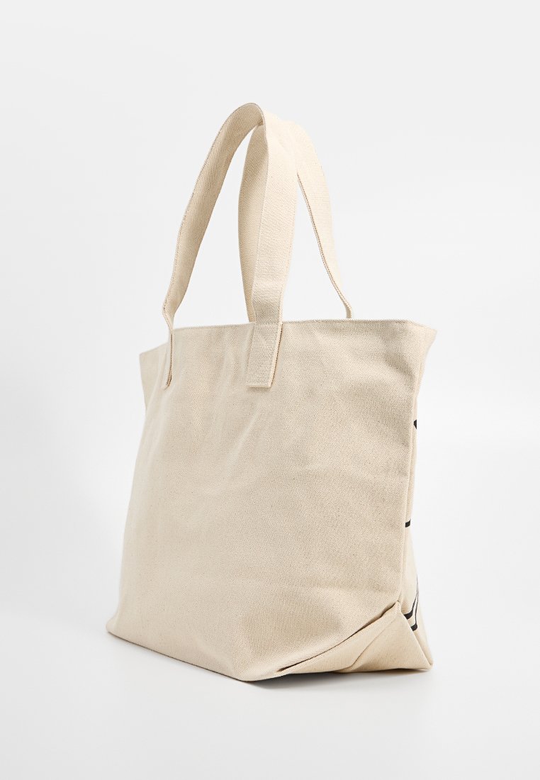 Sac en toile beige avec deux poignées, se tenant debout sur un fond blanc, texture de tissu légèrement froissée visible.